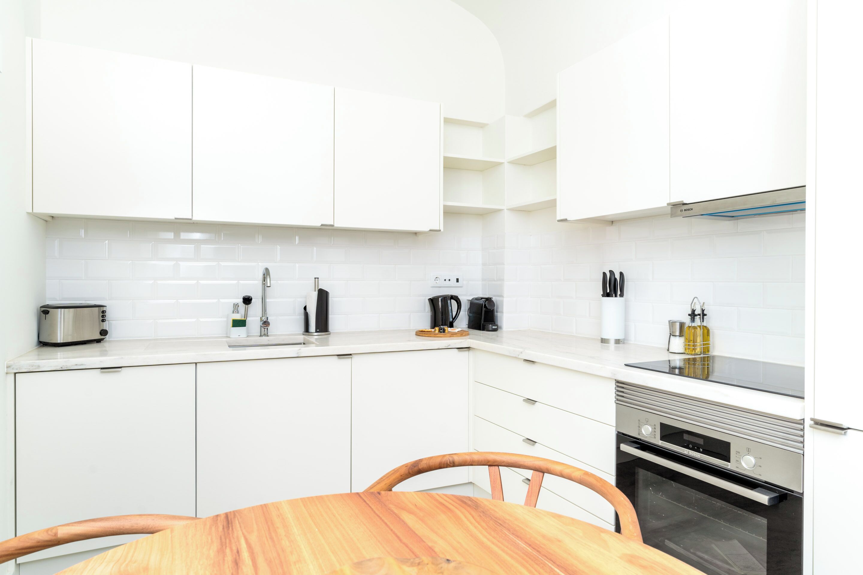 A modern kitchen with white cabinets, a wooden dining table, and a built-in oven.