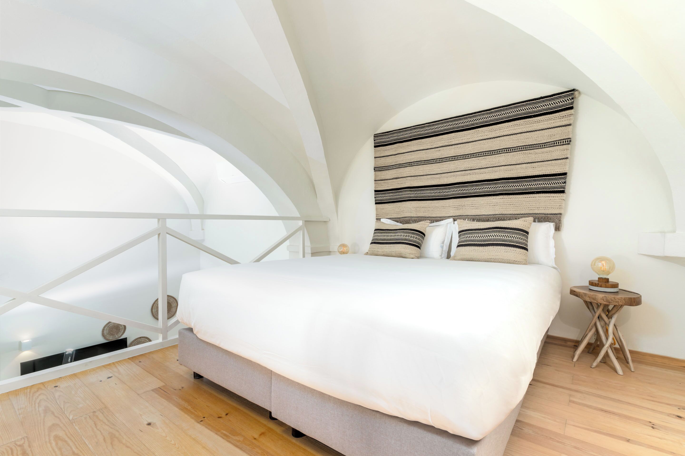 Une chambre avec un grand lit, un plancher en bois, un plafond blanc et une vue sur l’extérieur depuis un balcon vitré.