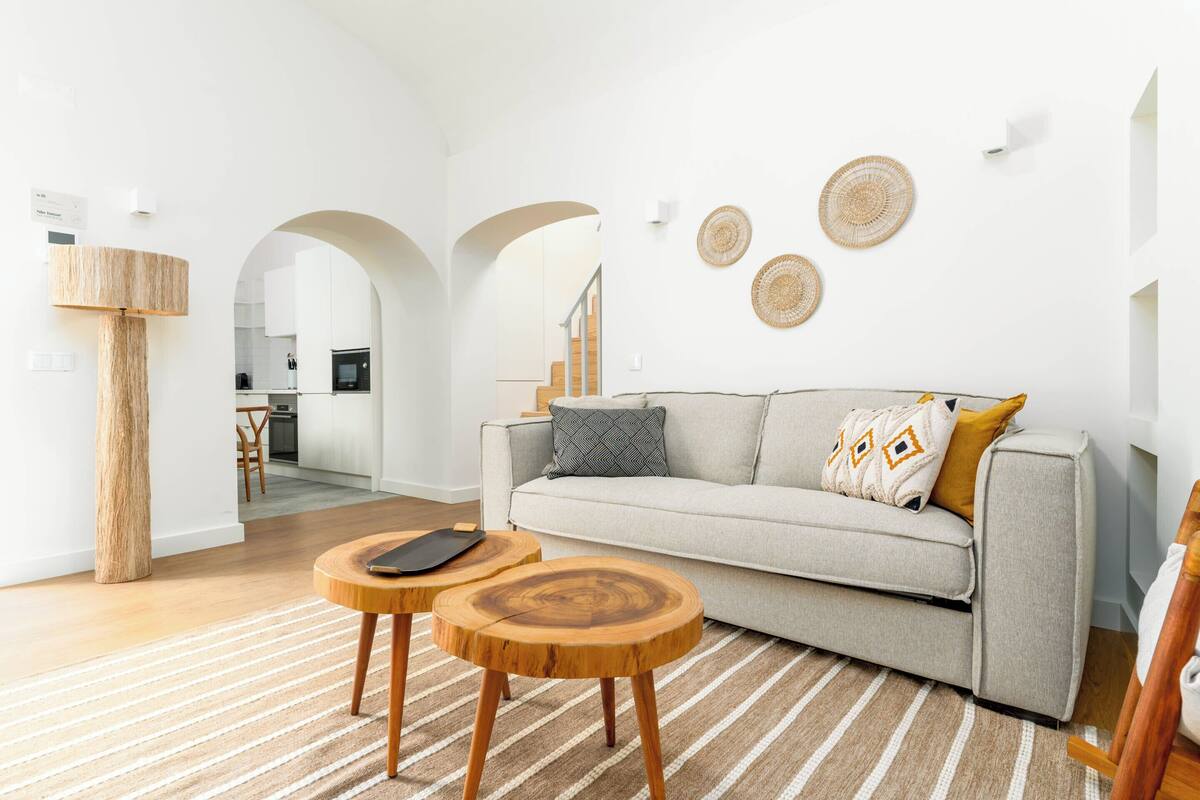 A modern living room with a sofa, two wooden side tables, a lamp, and a kitchen in the background.