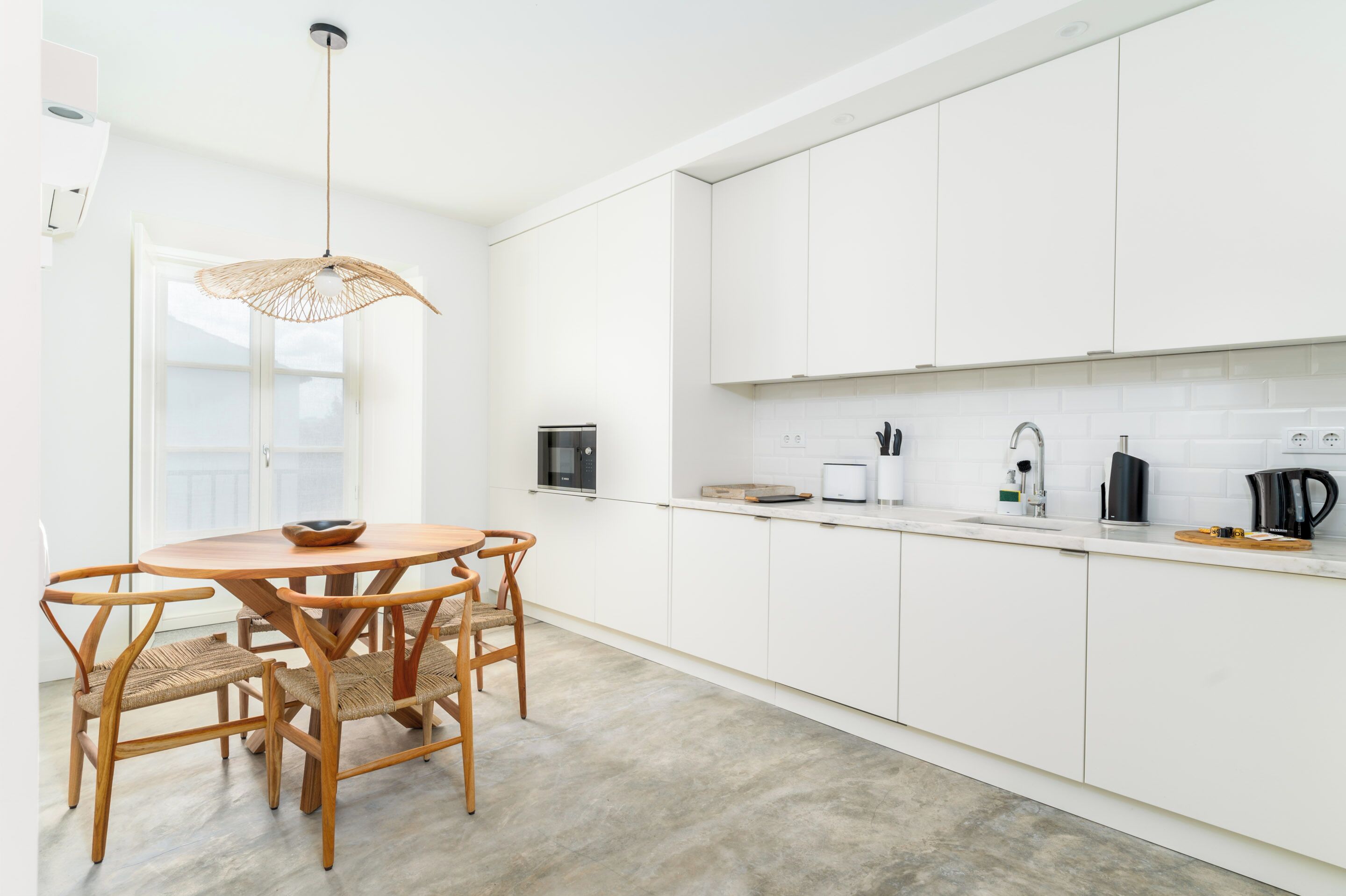 A modern kitchen with white cabinets, a round wooden table with chairs, a hanging light fixture, and a window with a view.