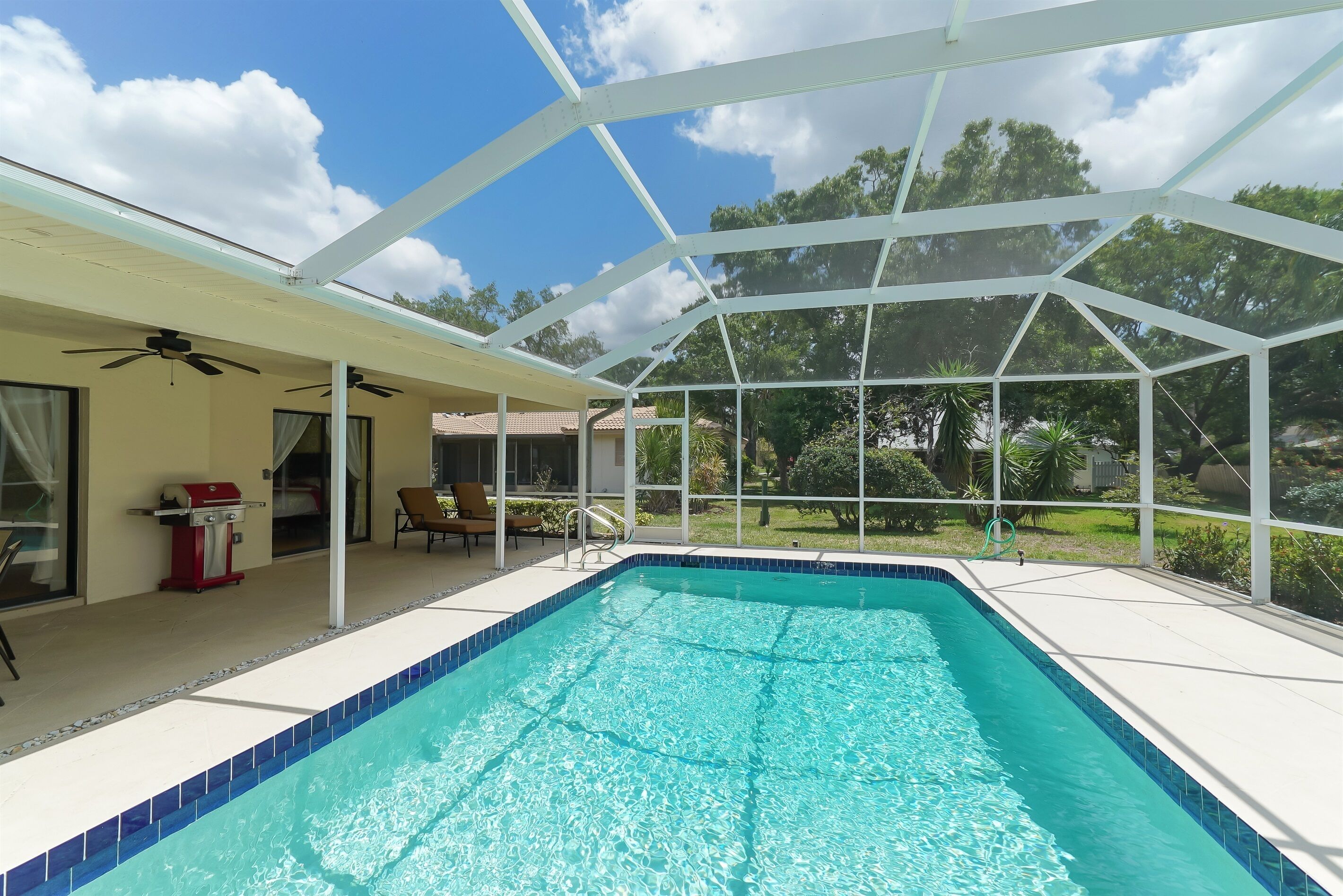 Une piscine dotée d’une structure en verre, d’un ventilateur de plafond et d’un espace extérieur aménagé avec des chaises et un barbecue.