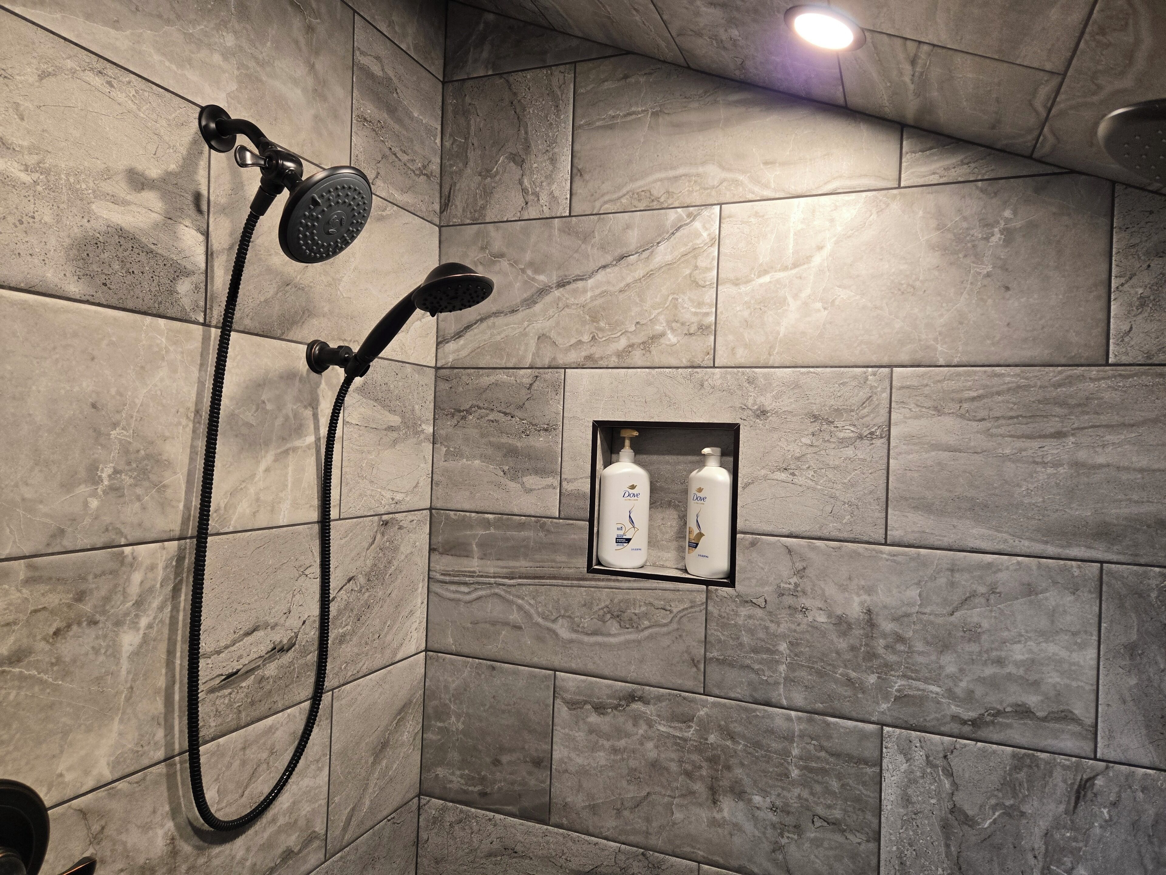 A shower area with a handheld showerhead and a wall-mounted showerhead, a small shelf with Dove shampoo bottles, and a recessed light above the shower.