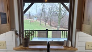 A kitchen with a double sink, wooden cabinets, a window with a view of a deck and trees, and a hanging light fixture. - Peaceful Meadows

 (Dundee)
