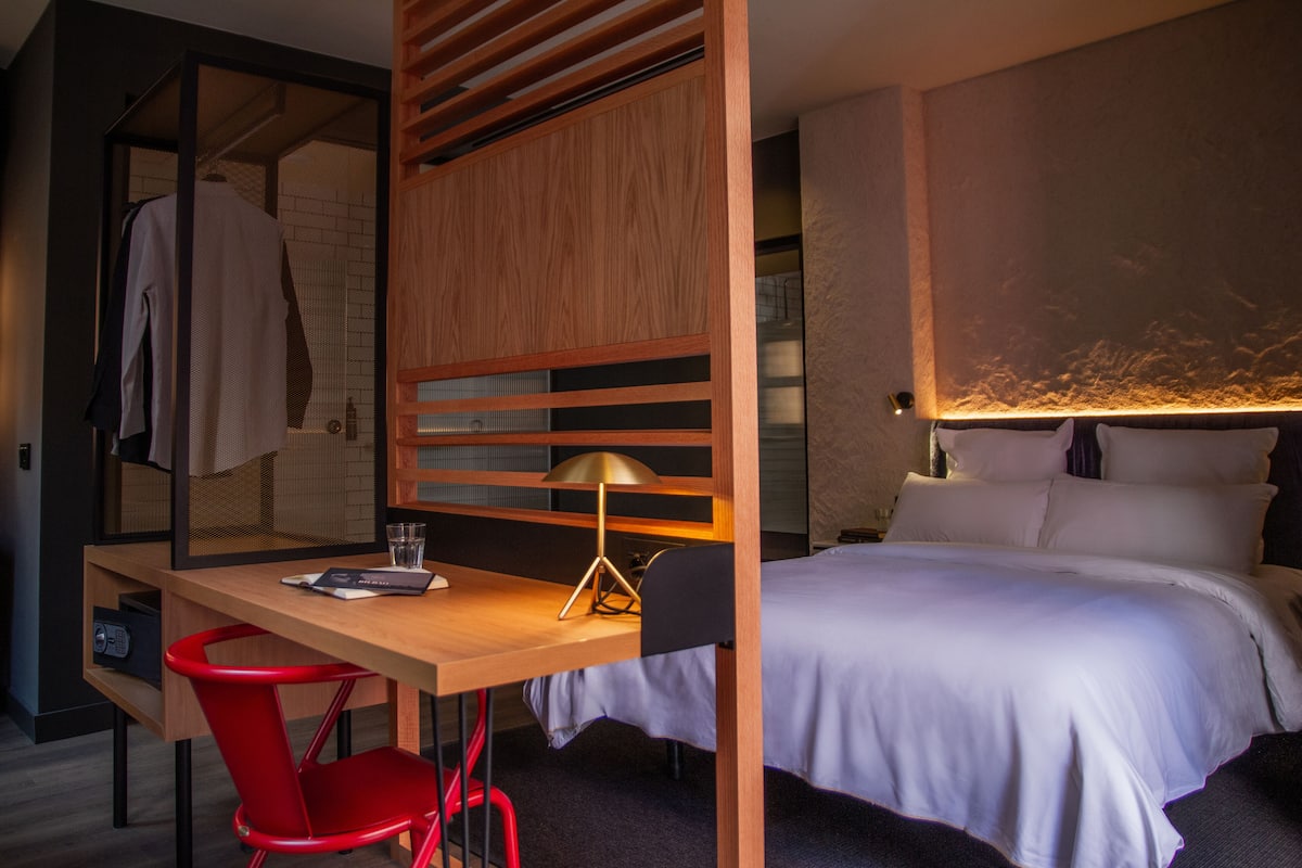 A hotel room with a wooden desk, a red chair, a bed with white bedding, and a lamp.