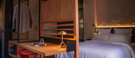 A hotel room with a wooden desk, a red chair, a bed with white bedding, and a lamp.