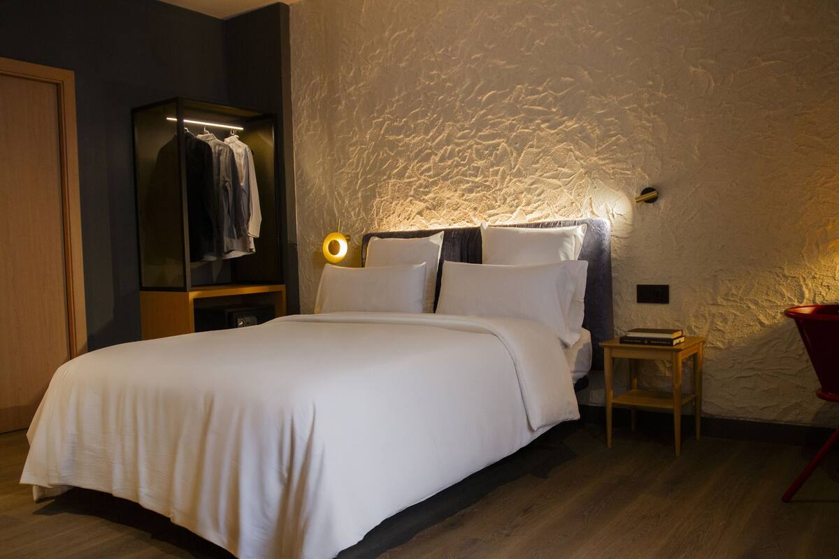 A neatly made bed in a room with a textured wall, a wooden nightstand, and a red chair.