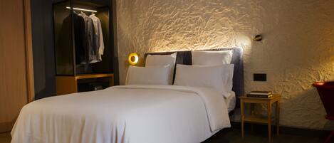 A neatly made bed in a room with a textured wall, a wooden nightstand, and a red chair.