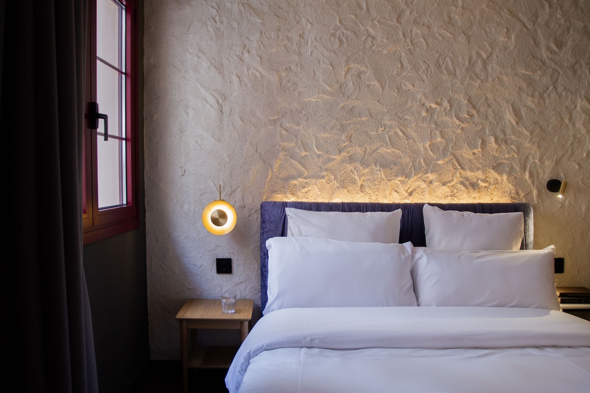 A bedroom with a textured wall, a bed with white bedding, a bedside table with a glass, and a wall-mounted lamp.