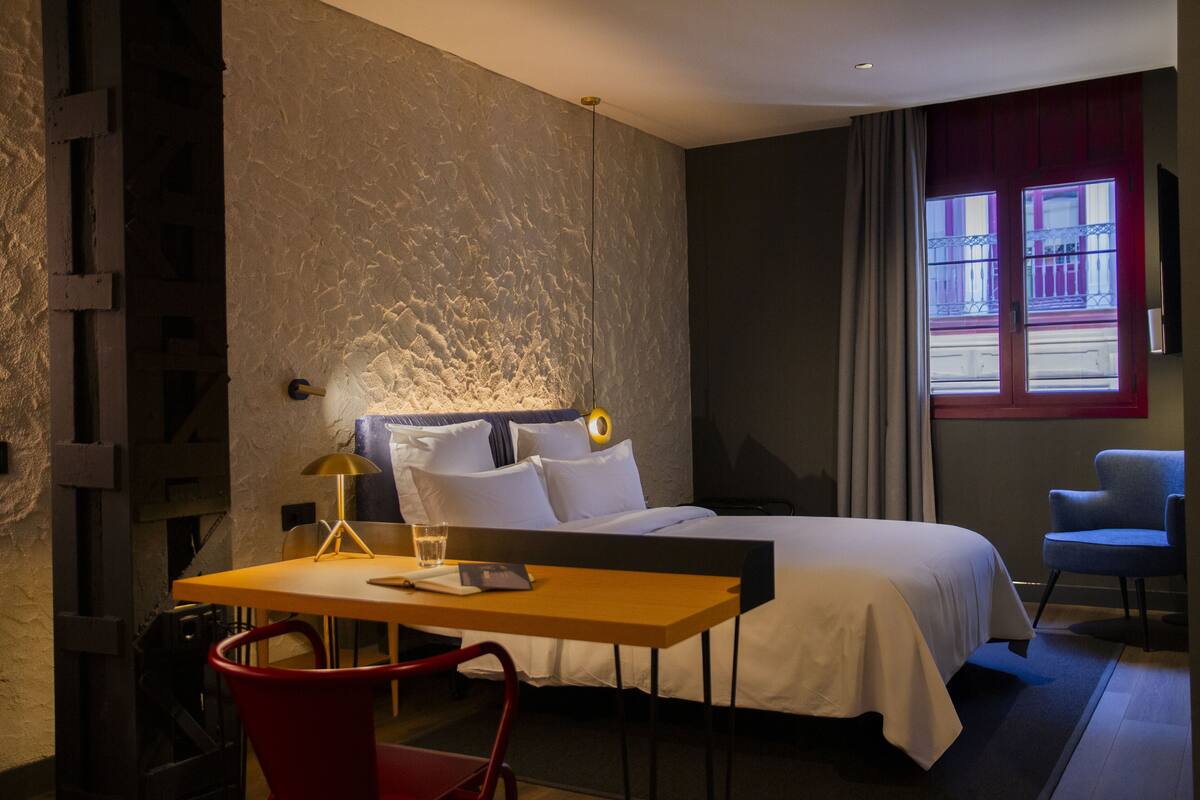 A hotel room with a bed, a desk, a chair, a lamp, and a window with curtains.