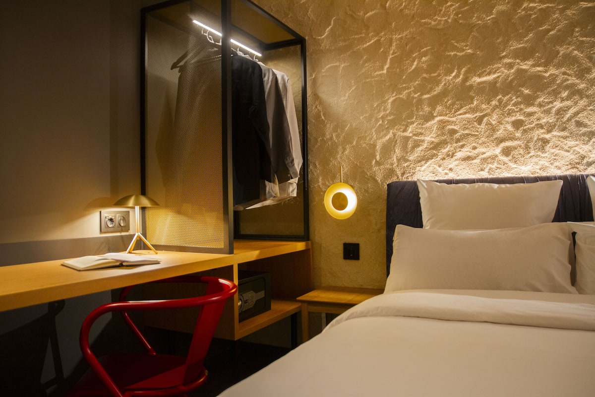A hotel room with a bed, a desk with a lamp, a red chair, a mirror, and a wall-mounted circular light.