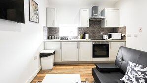 A compact kitchen with white cabinets, a microwave, and a stove. There is a black leather sofa with a patterned pillow and a small table with a white tablecloth. - Cosy Apartment Near Leeds City Centre (Leeds)