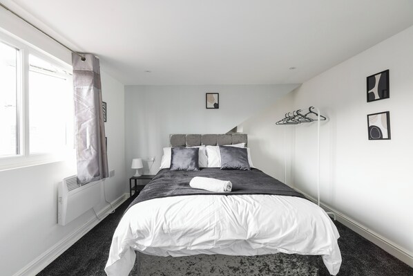 Una cama bien tendida con una colcha blanca y almohadas grises, un radiador, una ventana con cortinas y un perchero en la pared.