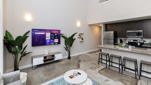 A modern living space with a flat-screen TV mounted on the wall, a kitchen area with stainless steel appliances, and a seating area with a round coffee table. - City Haven: Modern Loft 2bd/2bth Downtown DSM (Des Moines)