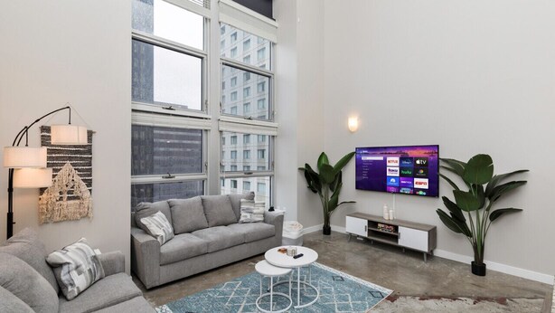 A modern living room with a grey sofa, a small white coffee table, a large window, and a flat-screen TV mounted on the wall. - City Haven: Modern Loft 2bd/2bth Downtown DSM (Des Moines)