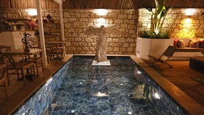 Una piscina interna con pareti in pietra, una statua al centro, arredi in legno e un soffitto in paglia.