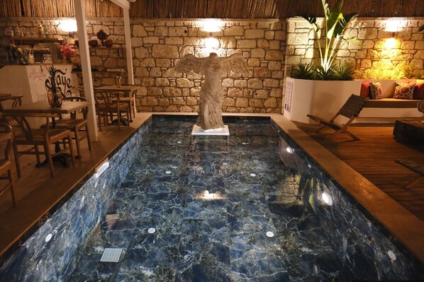 A stone-walled indoor pool with a statue in the center, wooden furniture, and a thatched ceiling. - Piraye Alaçatı (Cesme)