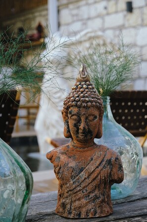 A Buddha statue in a rustic setting with a brick wall and potted plants in the background. - Piraye Alaçatı (Cesme)