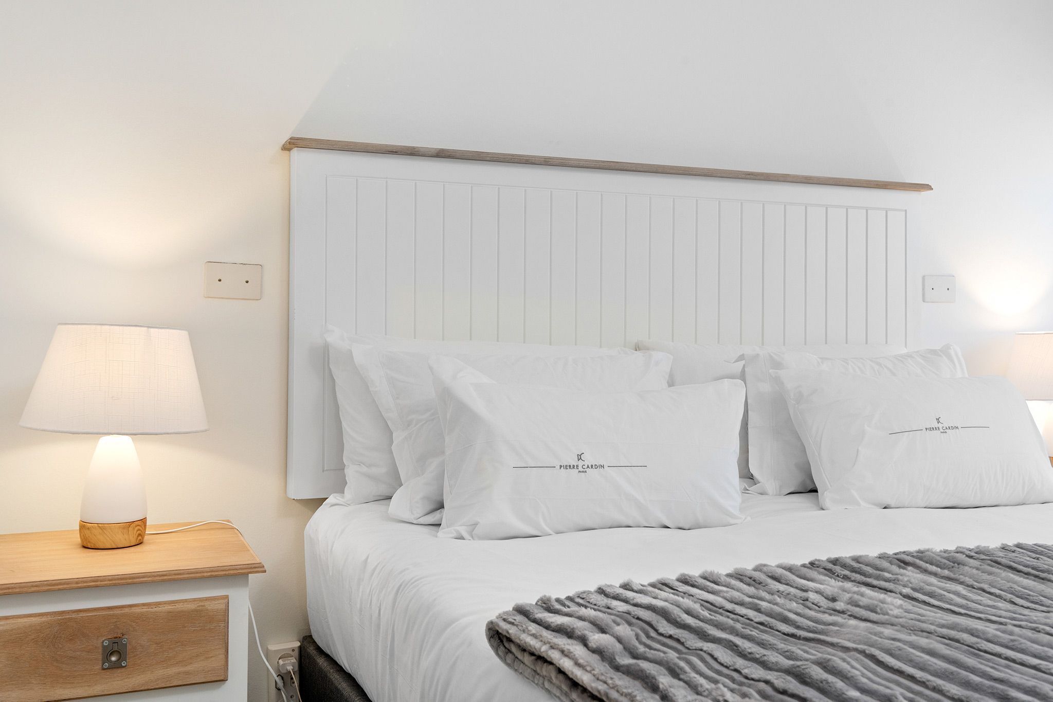 A neatly made bed with white bedding and pillows, a bedside lamp, and a wooden nightstand with a drawer.