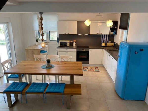 A modern kitchen with a dining area, featuring a wooden table, blue cushioned benches, a hanging light fixture, and a blue refrigerator.
