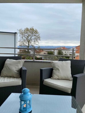 A balcony with wicker furniture and a table, overlooking a cityscape with buildings and mountains.