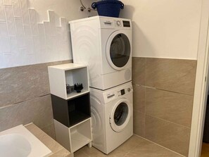 A compact laundry area with a washing machine and dryer stacked on top of each other, a small shelf unit, and a white sink.