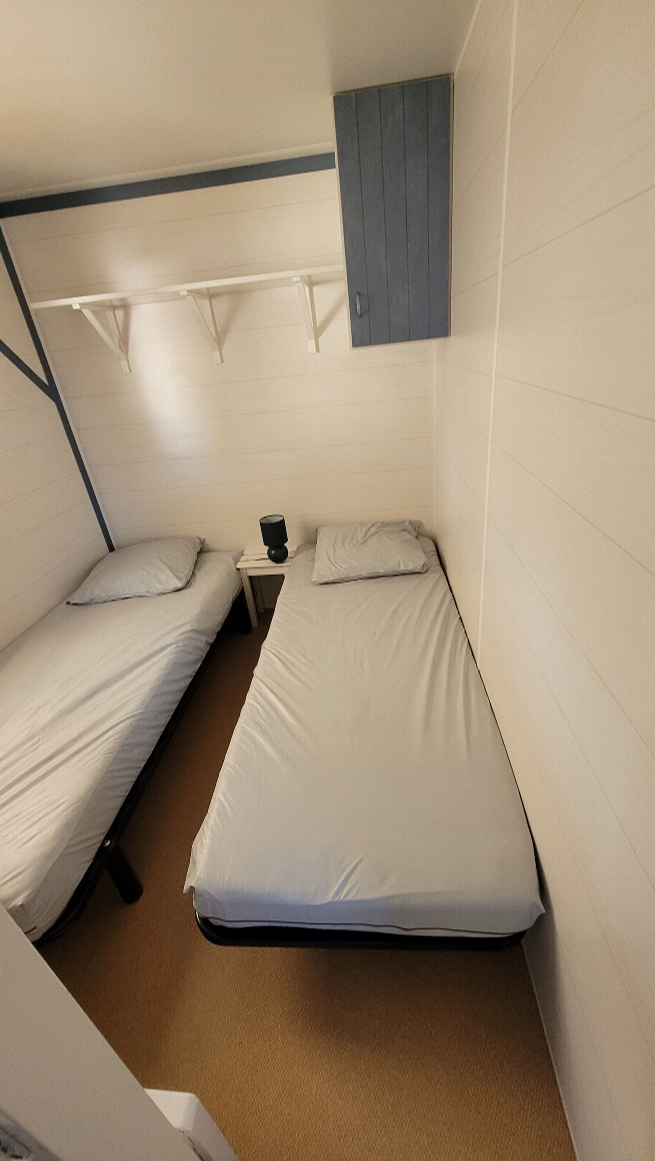 A small, compact room with two single beds, a small table with a lamp, and a cabinet above the beds.
