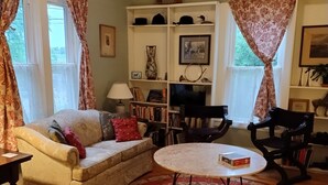 A living room with a sofa, a coffee table, a bookshelf, and a TV. - Quaint farmhouse with garden, big yard, river access- all near Middlebury, VT. (Weybridge)