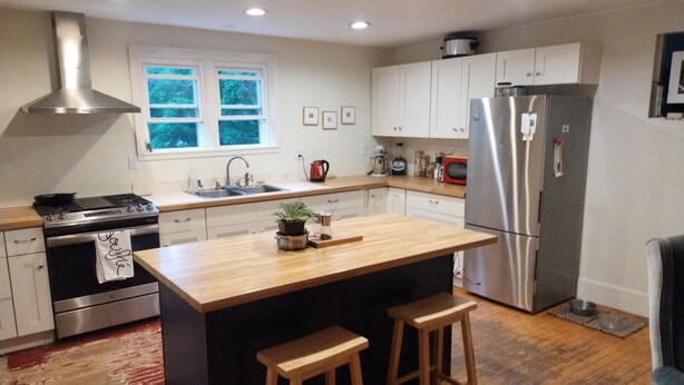A modern kitchen with white cabinets, stainless steel appliances, a large island with stools, and a window with a view of trees. - Quaint farmhouse with garden, big yard, river access- all near Middlebury, VT. (Weybridge)