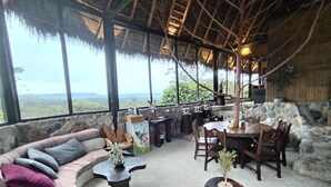 A cozy lounge area with a stone wall, a cushioned sofa, a round wooden table, and a dining set. - Finca de la Vaca - Casa el Mirador (Cumandá)