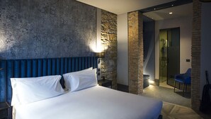 A hotel room with a large bed, a stone accent wall, a modern bathroom with a glass shower, and a blue chair. - Forvm Bijoux ROOMS (Trieste)