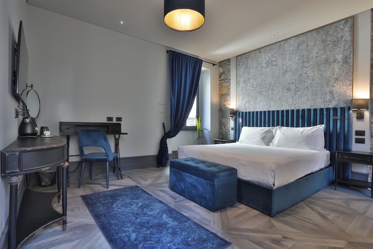 A modern hotel room with a large bed, a desk, a chair, and a blue ottoman.