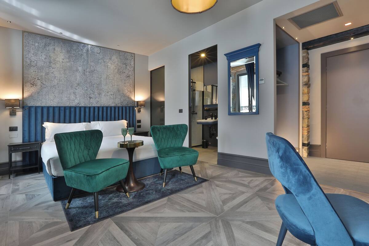 A modern hotel room with a large bed, two green armchairs, a small round table, and a large abstract painting on the wall.