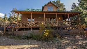 A two-story log cabin with a green roof, a covered porch, and a stack of firewood in front. - Caja de Tesoro ~ A Romantic Mountain Getaway! (Arroyo Seco)