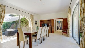 Une salle à manger spacieuse avec une grande table et des chaises, de larges fenêtres avec des rideaux et une vue sur la verdure environnante.