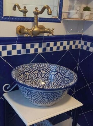 Een badkamer met een blauw-wit geruit betegelde achterwand, een gouden kraan, en een decoratieve blauw-witte kom op een witte plank.