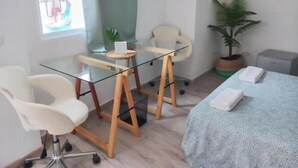 A modern room with a glass desk, two white chairs, a bed with a blue cover, and a potted plant. - Guesthouse Jazmines Mozàrabes (Baena)