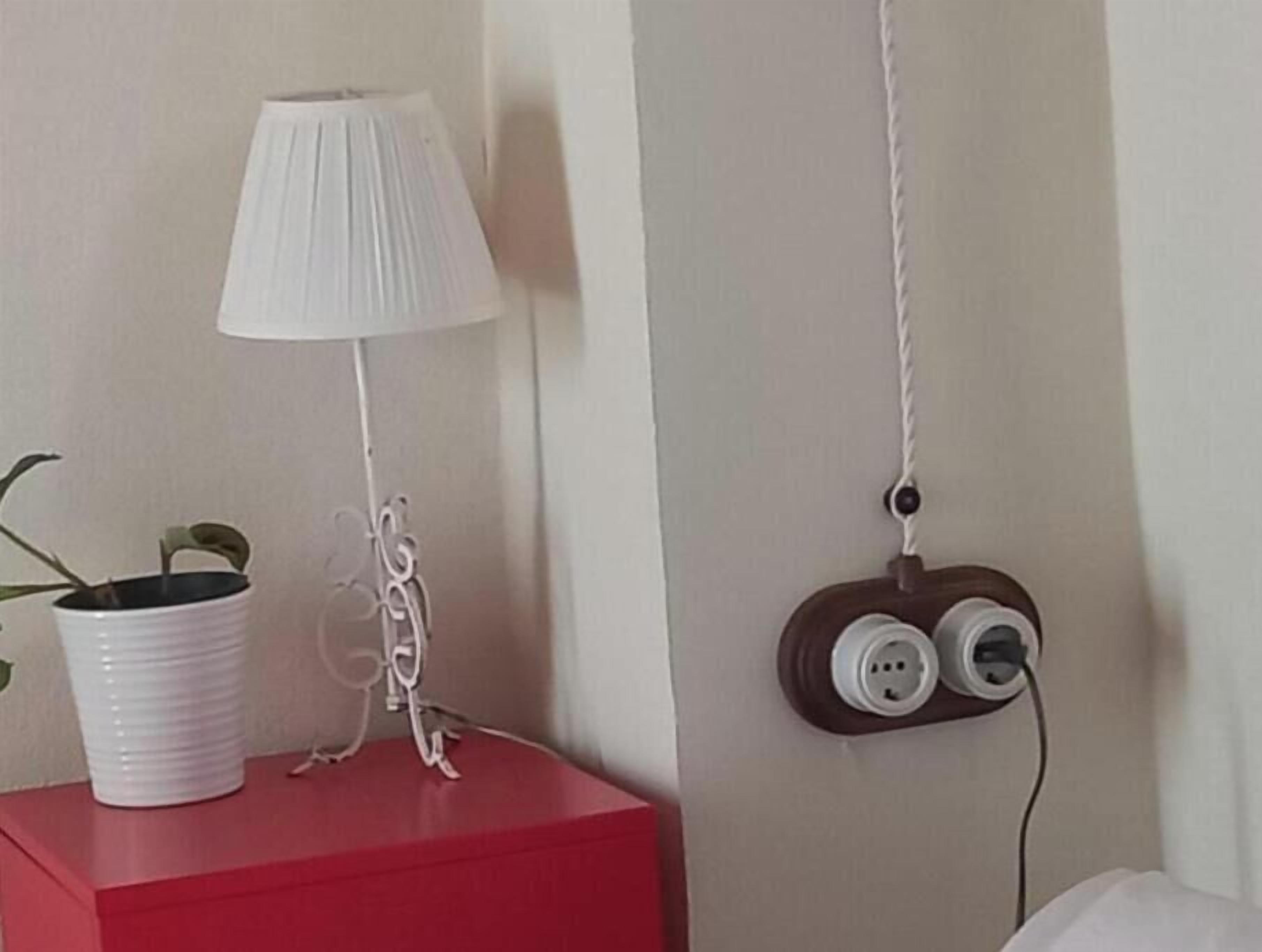 Une table de chevet rouge, sur laquelle se trouve une lampe blanche et un pot de fleurs.
