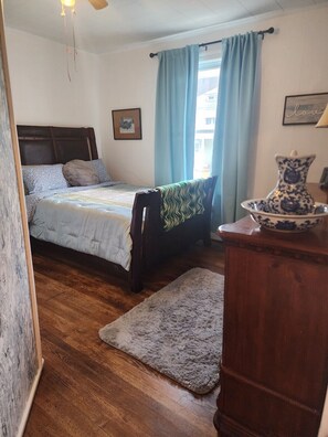 Ein Schlafzimmer mit einem Bett, einem Holz-Kopfende, einem Nachttisch, einer Vase und einem Fenster mit Vorhängen.