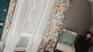 Une pièce avec une chaise blanche, un rideau à motifs floraux et une vue sur un bâtiment à l’extérieur.