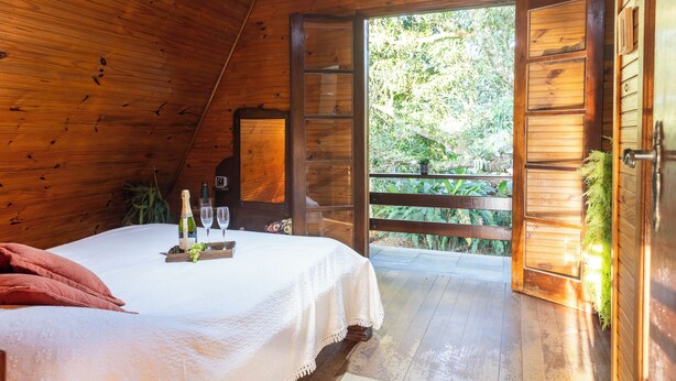 A wooden room with a bed, a table with a bottle and glasses, and a door leading outside. - Natureza-piscina-lareira-Brazilan Corner (Aruja)