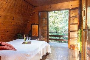 Quarto de madeira com uma cama, uma mesa com uma garrafa e copos, e uma porta que dá para o exterior.