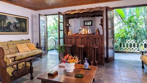 A cozy living area with a wooden ceiling, a sofa, a coffee table with a floral centerpiece, and a bar area with a view of lush greenery. - Natureza-piscina-lareira-Brazilan Corner (Aruja)