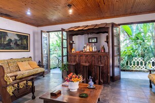 Uma sala de estar acolhedora com teto de madeira, um sofá, uma mesa de centro com um arranjo floral e um bar com vista para o verde exuberante.