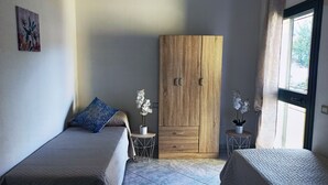 A bedroom with two beds, a wooden wardrobe, and a window with curtains. - Welcomely - Casa San Pietro (Budoni)