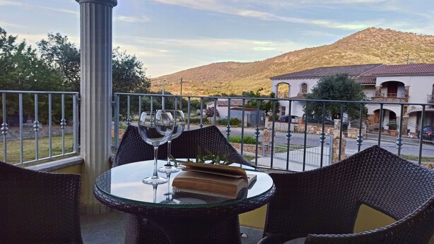 A balcony with wicker chairs and a glass table, overlooking a street and a hill. - Welcomely - Casa San Pietro (Budoni)