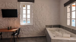 A bathroom with a stone wall, a bathtub filled with bubbles, a window, and a flat-screen TV mounted on the wall. - Horizon 4 You Mykonos (Mykonos)