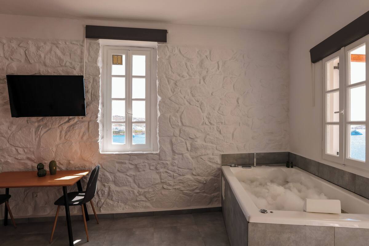 A bathroom with a stone wall, a bathtub filled with bubbles, a window, and a flat-screen TV mounted on the wall.