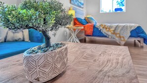 A room with a bonsai tree in a geometric pot, a blue sofa, a bed with a colorful blanket, and a painting on the wall.
