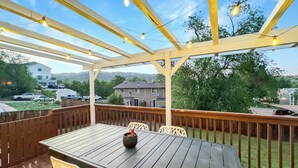 A wooden deck with a table and chairs, string lights, and a view of houses and trees in the background.