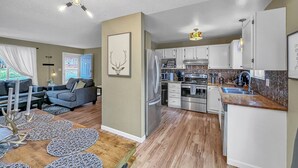A modern kitchen with stainless steel appliances, a dining area with a wooden table and patterned placemats, and a living room with a gray sofa and a decorative wall piece.